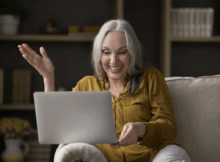 Smiling Fortyish woman with long straight white hair comfortably on a sofa and playing online casino games on her laptop
