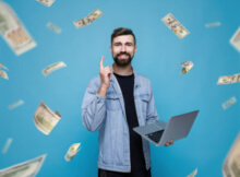 smiling young bearded man standing with laptop as money floats all around him symbolizing great online casino bonuses