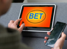 Man with tablet BET in large blue letters and orange and yellow background. Also holding a smartphone for online casino gaming.