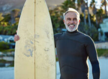 older man with short salt and pepper beard with big smile holding surfboard indicating good life in Australia