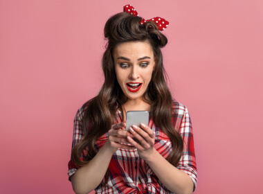 young woman in 1950's hairdo with red hair ribbon with white polka dots happily playing casino games on her smartphone