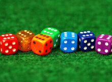 seven dice all of a different color laid out on a green felt table all showing the number six