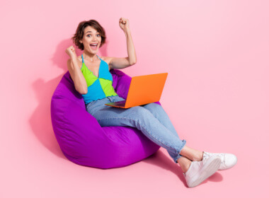 happy young woman playing online casino games sitting on large purple floor pillow