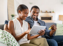 attractive young couple using a credit card to make a payment online on their tablet. They are sitting on their sofa at home.