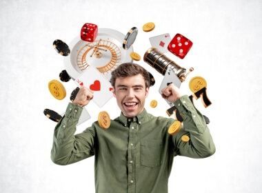happy young man raising arms after winning at an online casino. Casino props are around him: chips, dice, roulette wheel etc