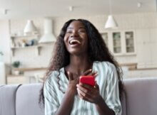 Ecstatically happy young black woman on the sofa with her phone celebrating a bonus from an online casino.
