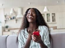 Ecstatically happy young black woman on the sofa with her phone celebrating a bonus from an online casino.