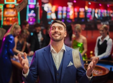 A man enjoying himself immensely at a land-based casino. He has a blue suit and vest with a short beard and mustache and no tie.